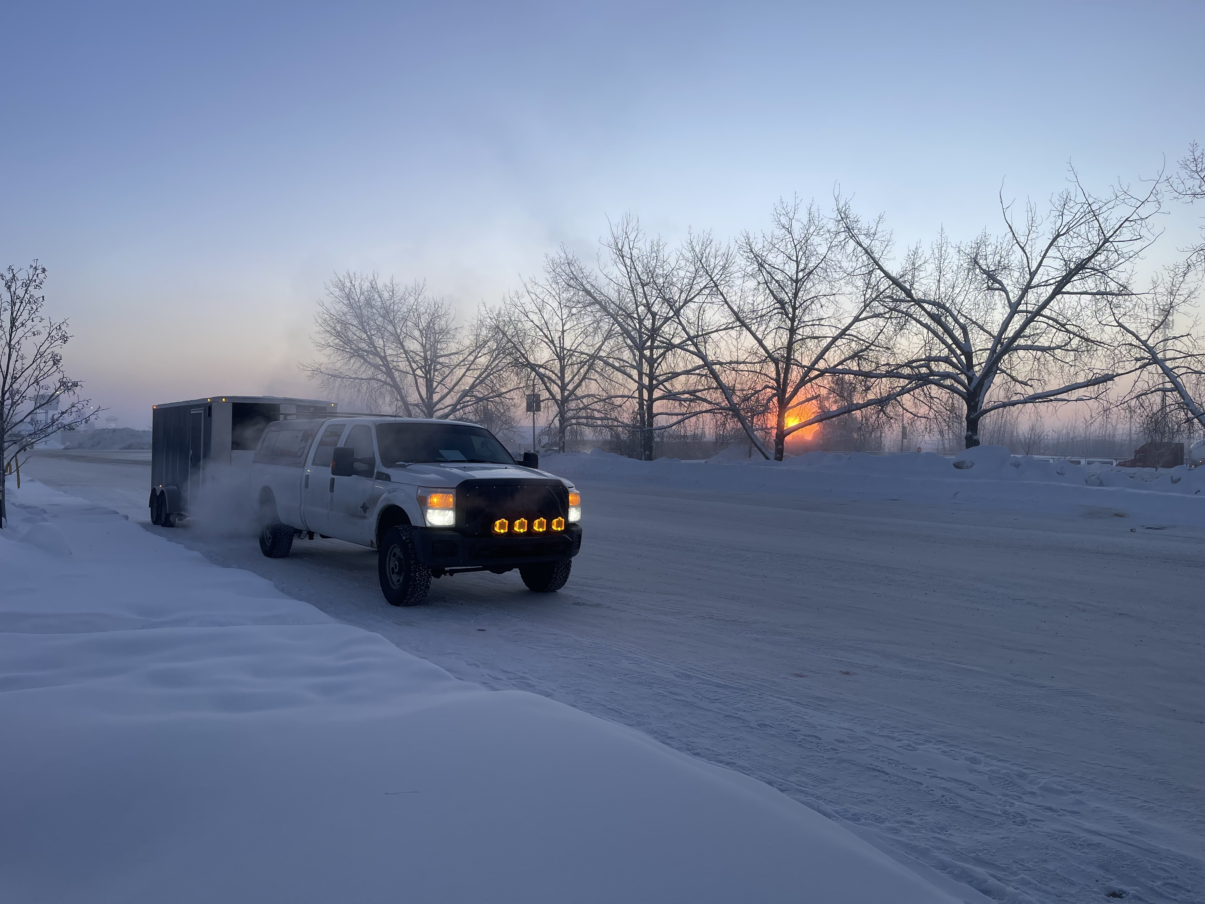 White pickup towing an enclosed trailer down a snowy Maine road at sunrise